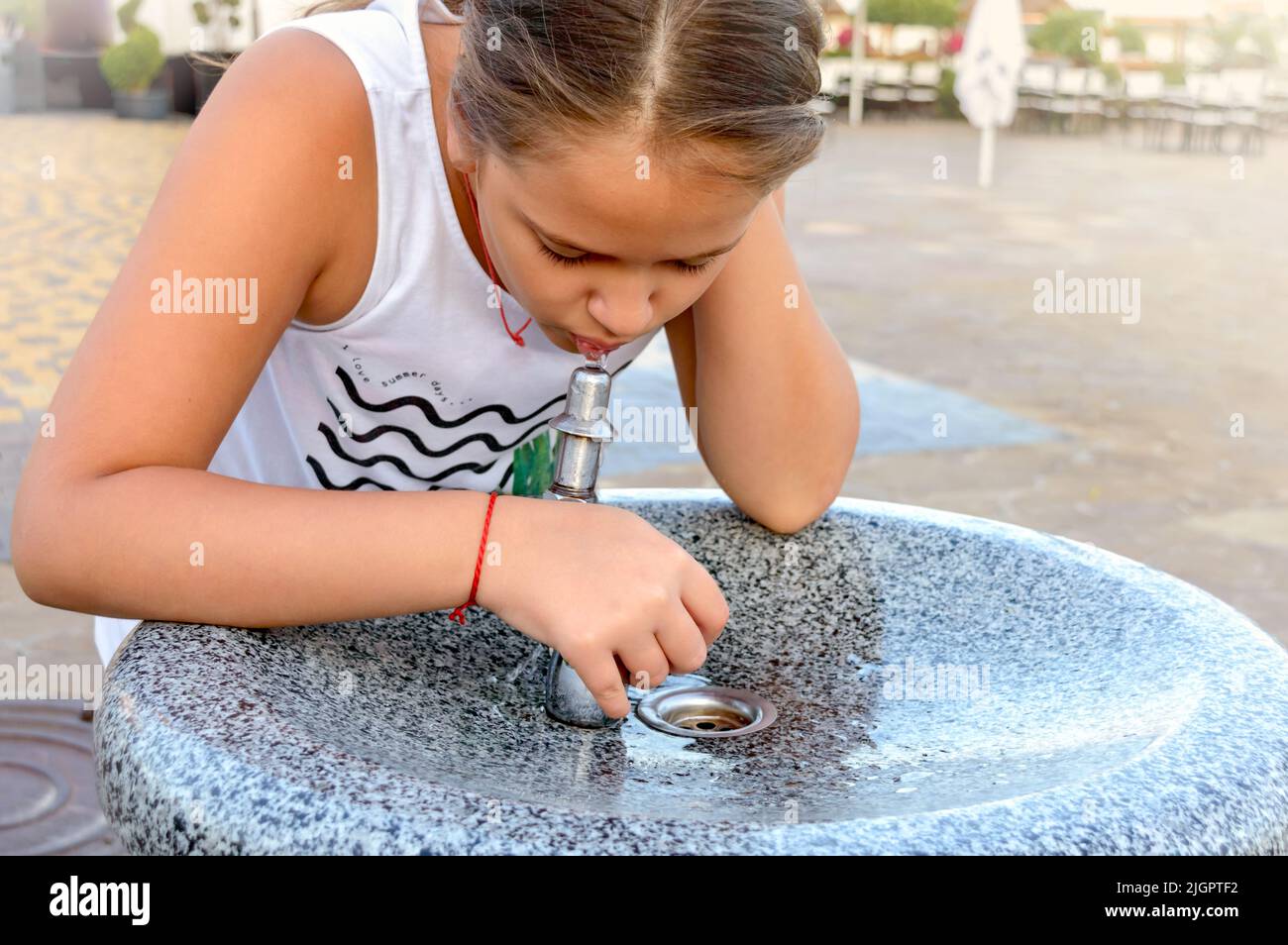 Cute little girl drinks water from public potable fountain faucet fountain on a hot summer day. Stock Photo