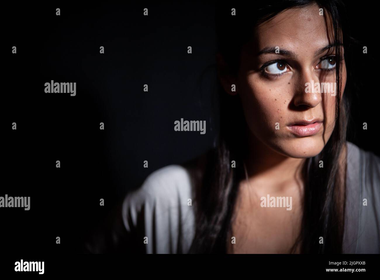 Dark Fear. A concerned look on the face of a beautiful young mixed-race Anglo-Indian female model in a dark place. From a series with the same model. Stock Photo