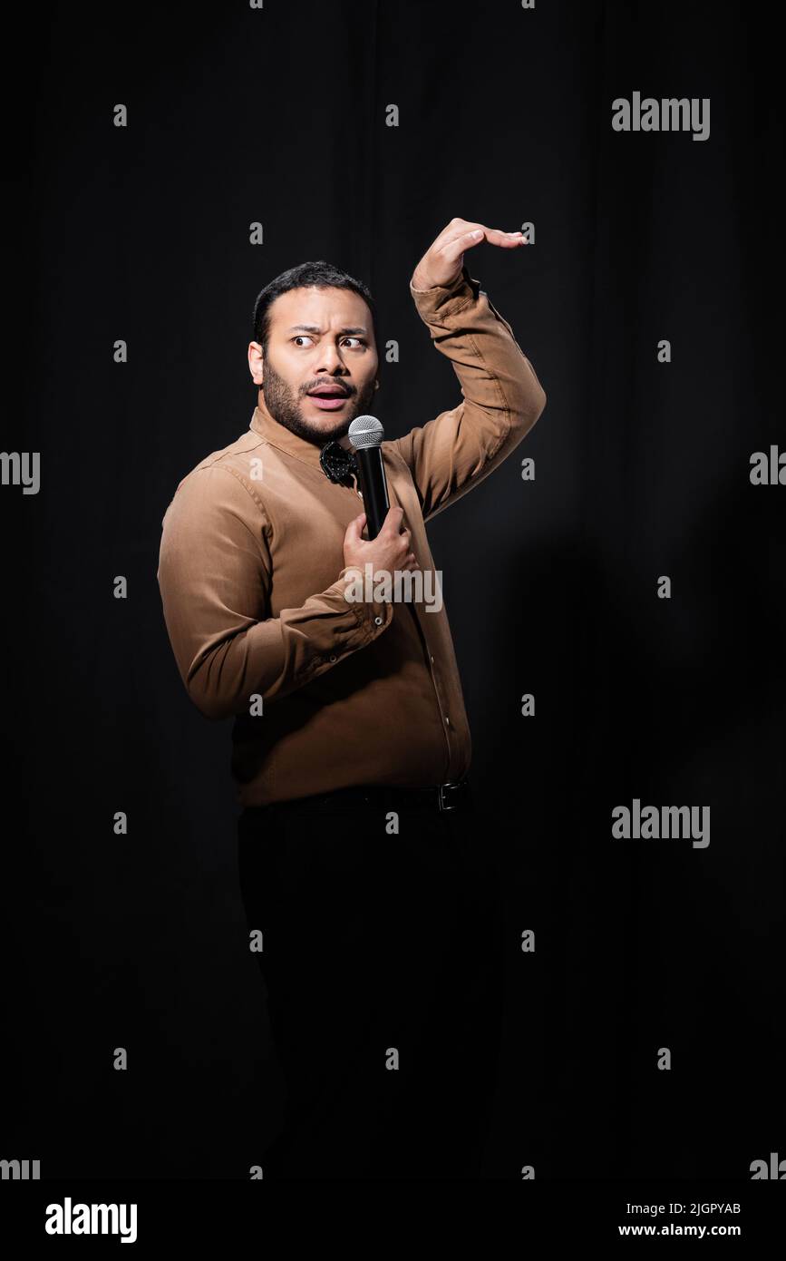 indian stand up comedian gesturing while telling joke into microphone on dark stage on black Stock Photo