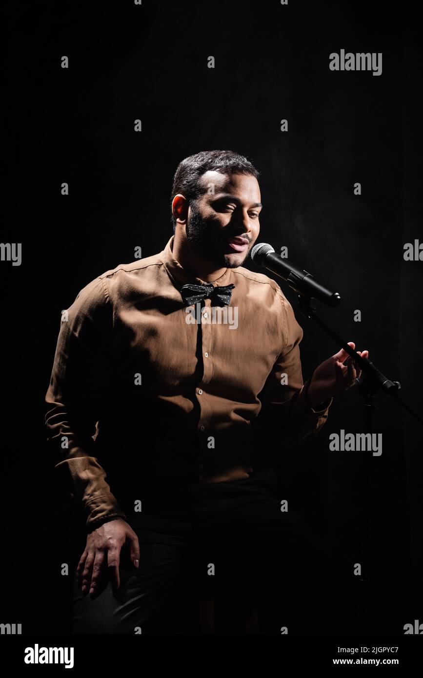 indian stand up comedian telling jokes into microphone on dark stage isolated on black Stock Photo