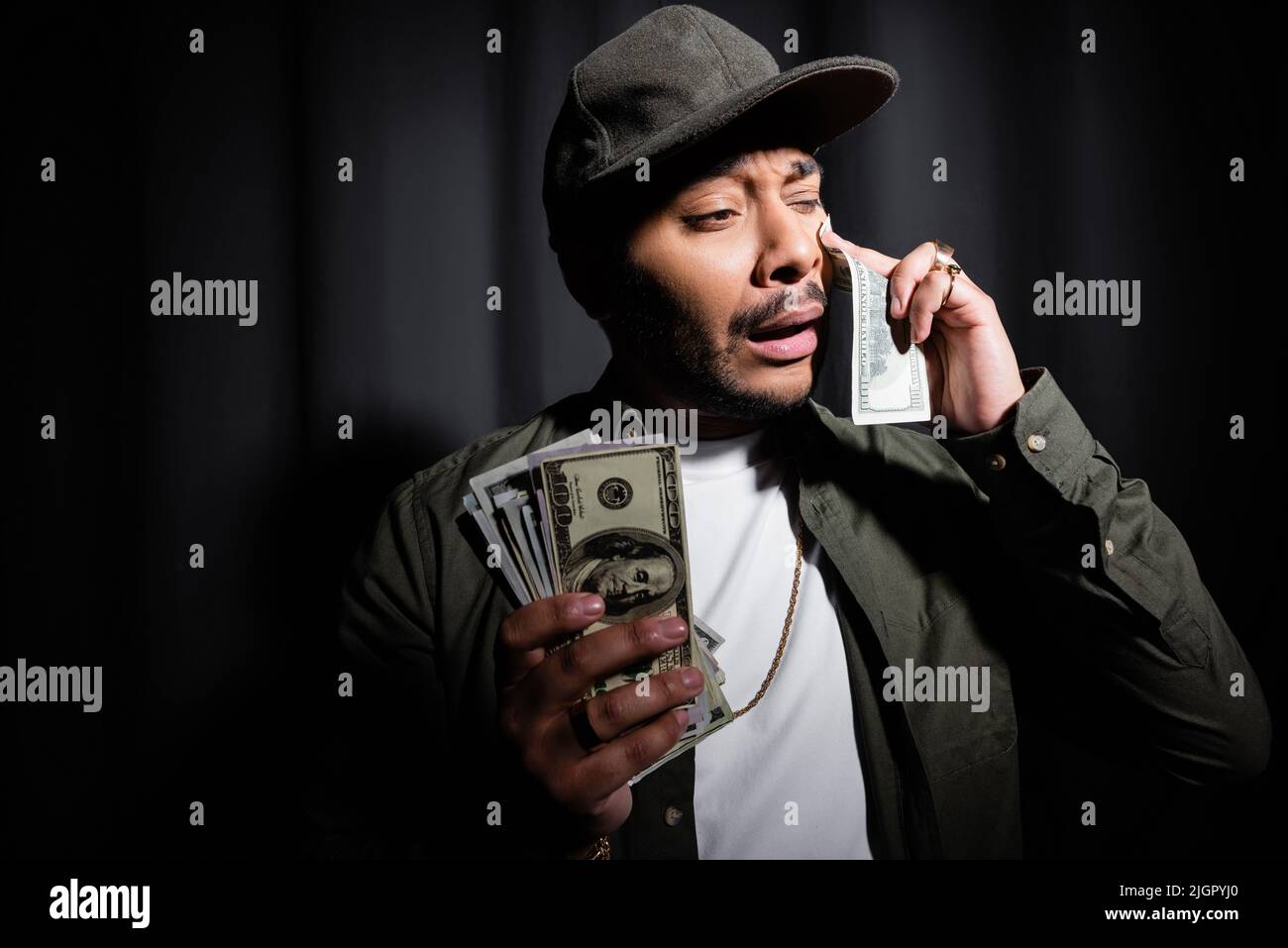 sad indian hip hop performer in cap crying and wiping tear with dollar banknote on black Stock Photo