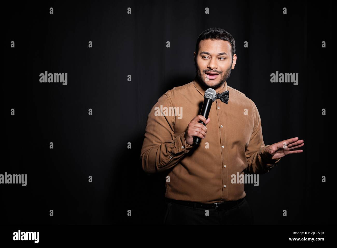 indian stand up comedian with bow tie holding microphone during monologue on black Stock Photo