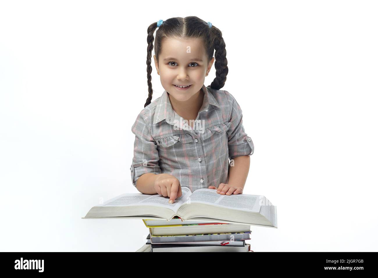 charming little girl with braided pigtails sits in front of a large book and looks into the camera. Stock Photo