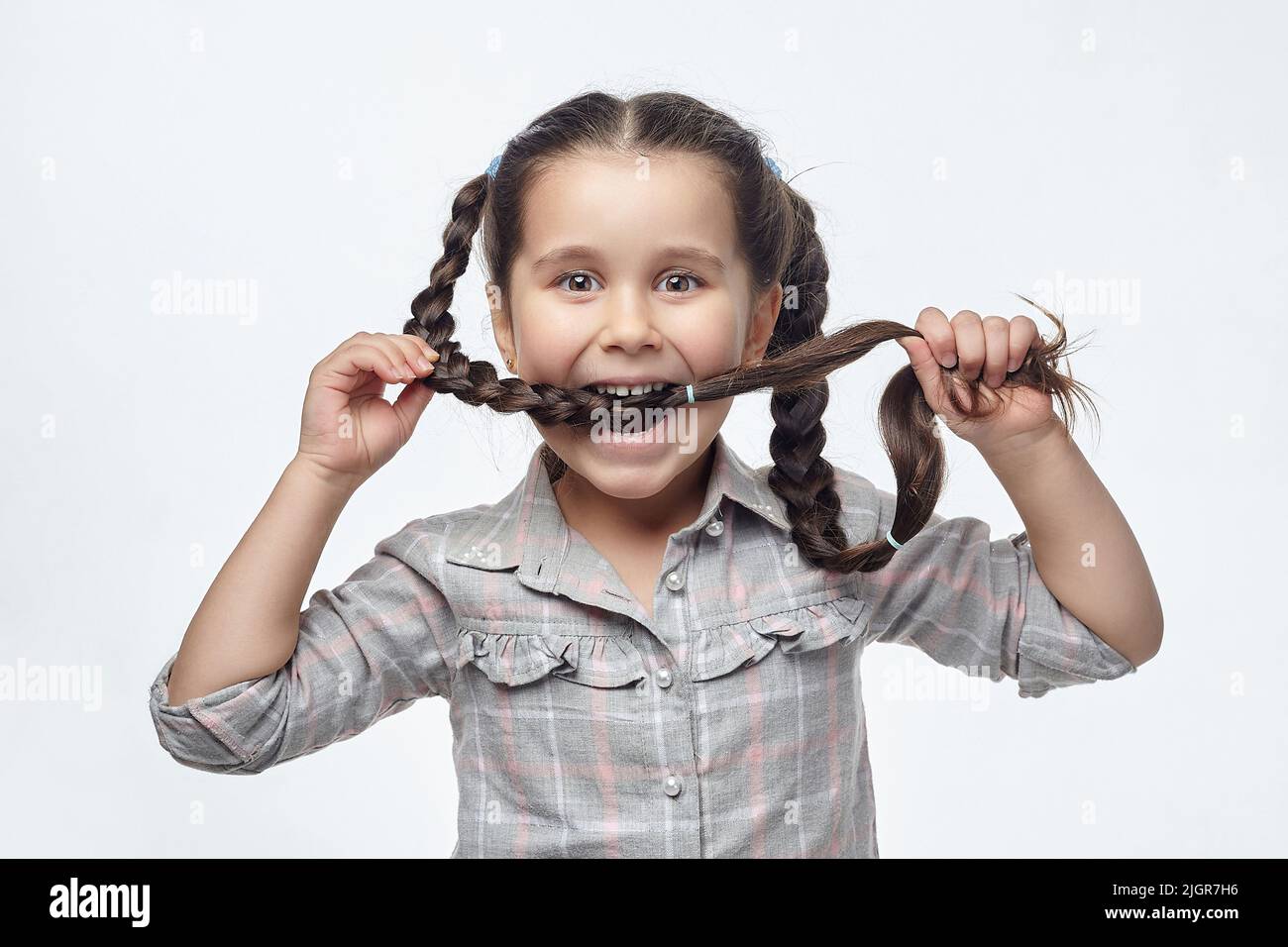 portrait of a beautiful little black-haired girl in close-up. she bites her braided pigtail with a smile. Stock Photo