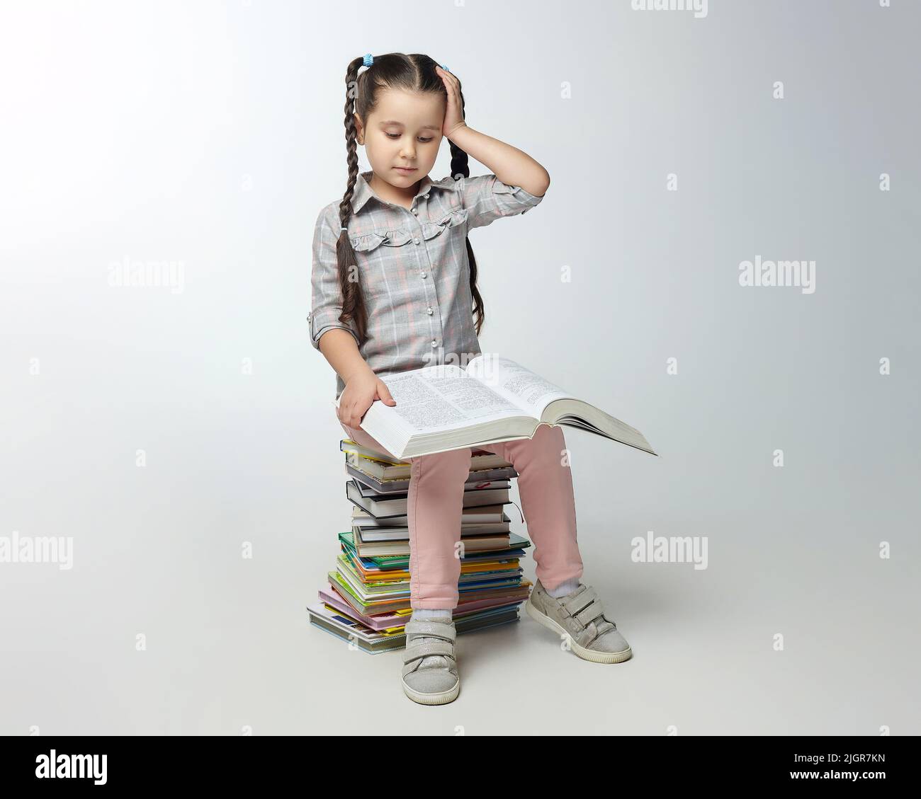 little girl with braided pigtails sits on a large stack of books and ...