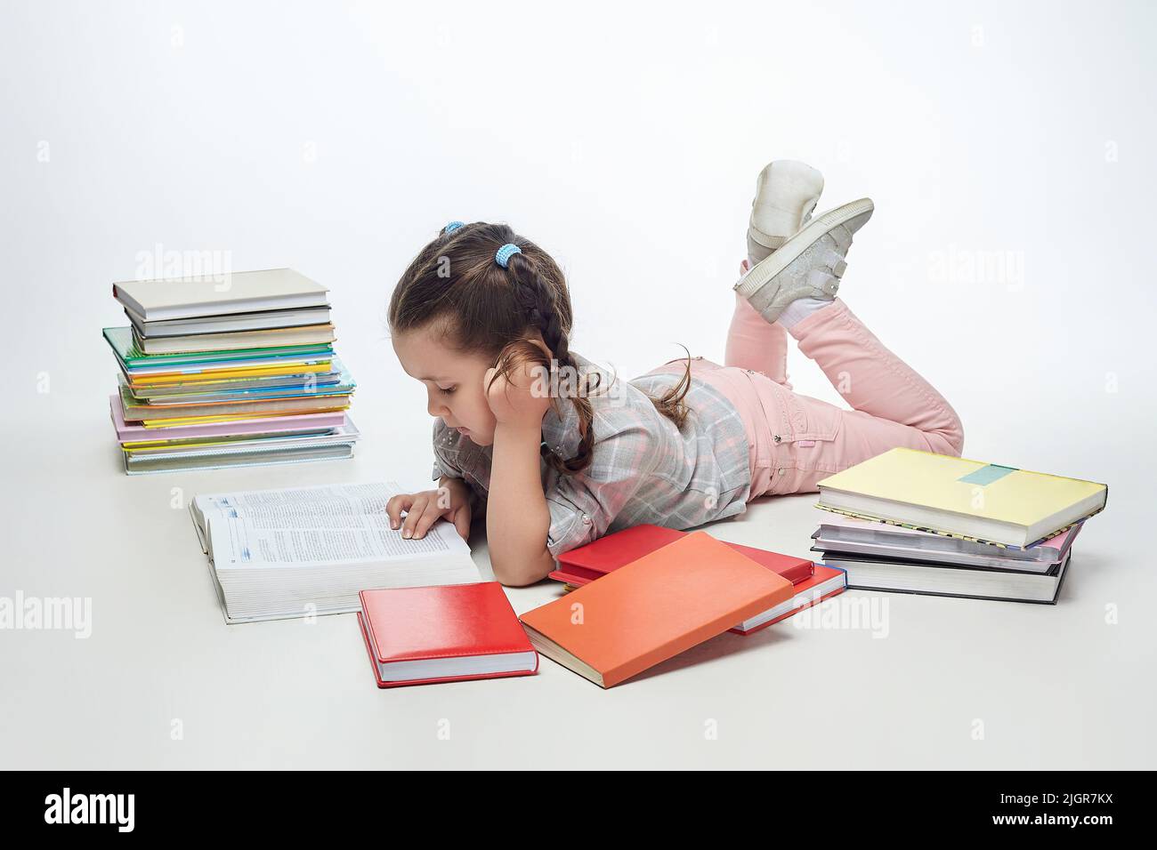 charming little girl with braided pigtails in the studio on a white background, a lot of books are scattered around her. Stock Photo