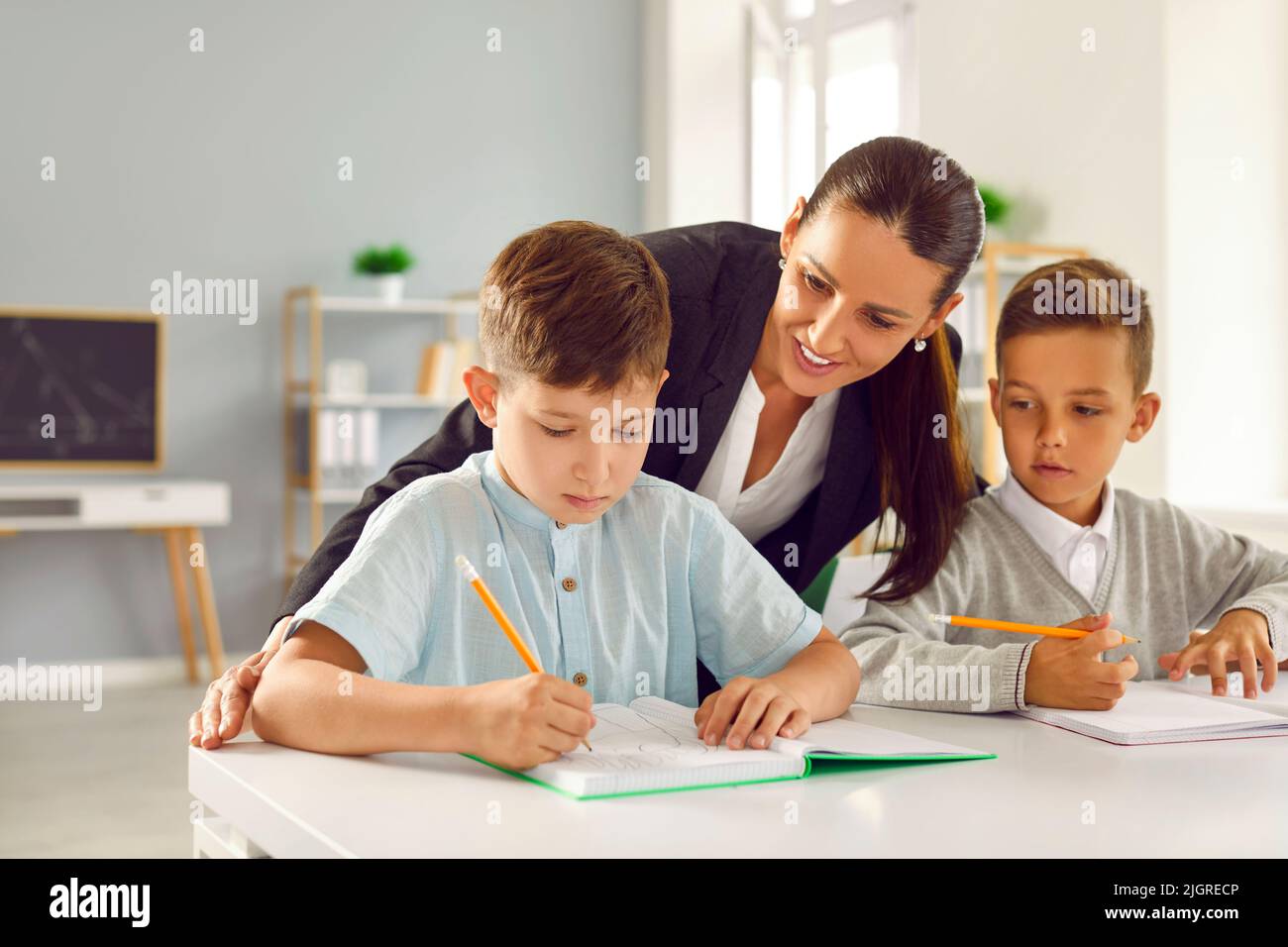 Friendly teacher helps small schoolchildren with test tasks in elementary school classroom. Stock Photo