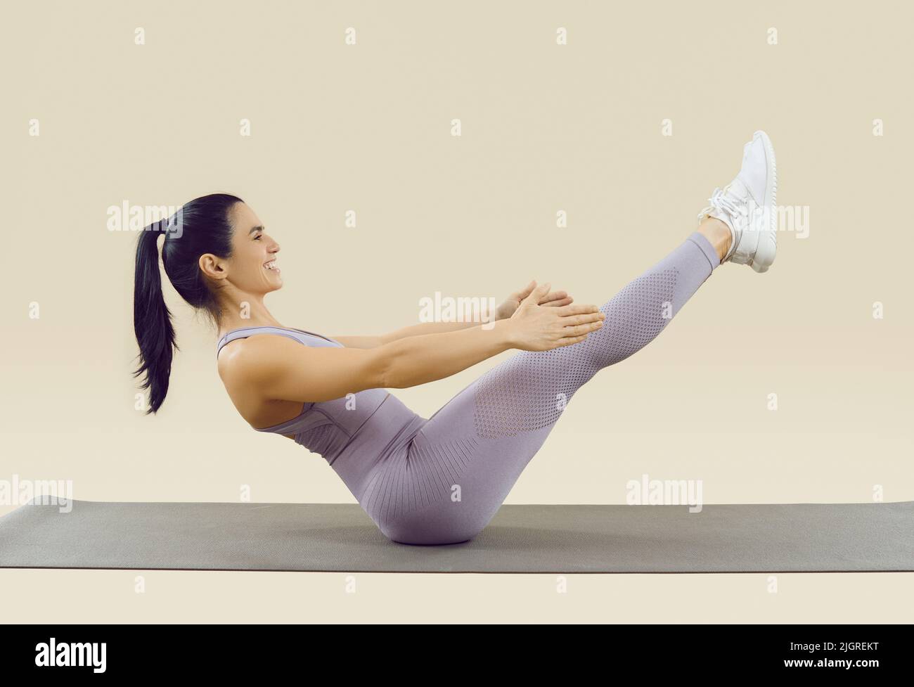 Smiling sporty woman in sportswear doing abs exercise with leg raise on beige background. Stock Photo