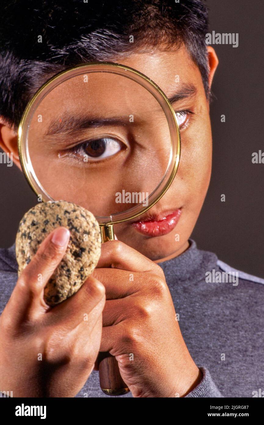His eye enlarged by a magnifying glass, an Asian American boy examines a geological sample stone. Stock Photo