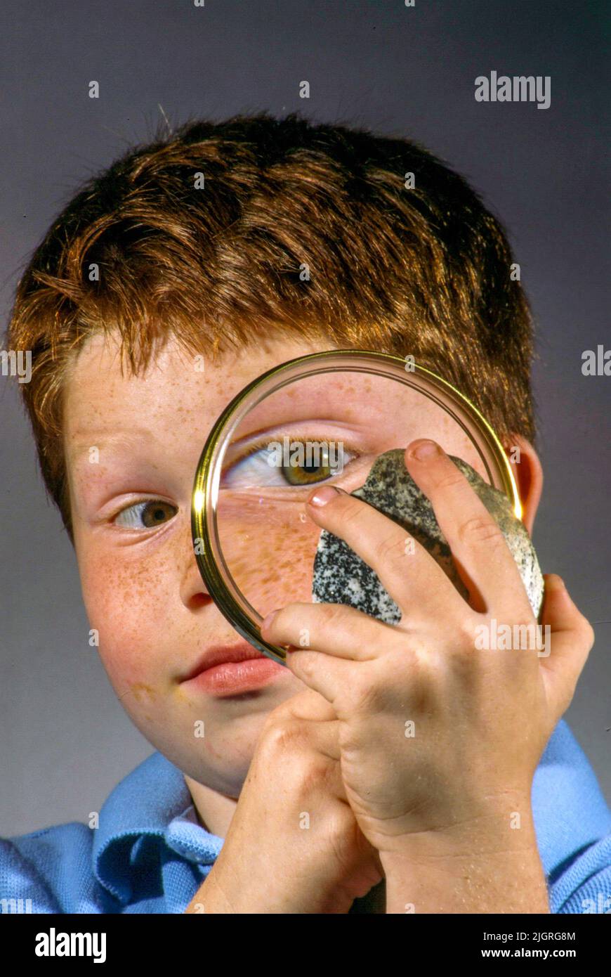 His eye enlarged by a magnifying glass, a red headed boy examines a geological sample stone. Stock Photo