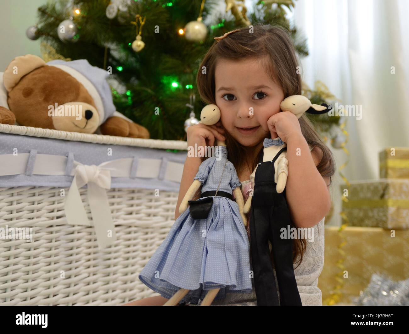A girl sits against the background of a Christmas tree and a basket with a bear's head in a blue hat in her hands. She holds 2 deer, a boy and a girl, she leans against their cheek and looks Stock Photo