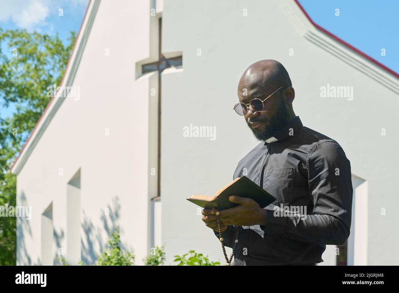 Young serious black man in shirt with clerical collar and eyeglasses reading Holy Bible while praying or preaching by church building Stock Photo
