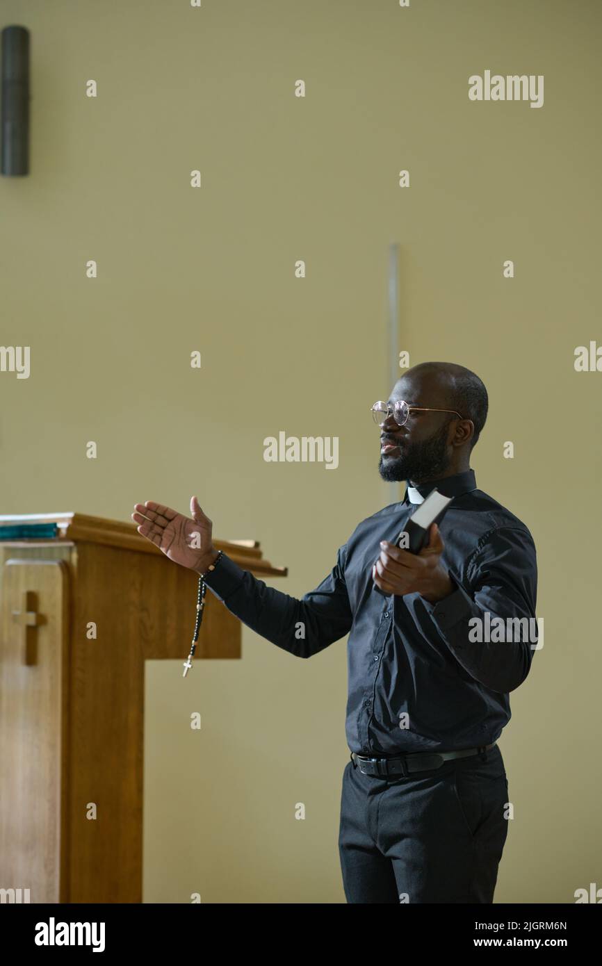 Young confident pastor in black shirt with clerical collar and trousers blessing pashioners while holding rosary beads and Holy Bible Stock Photo