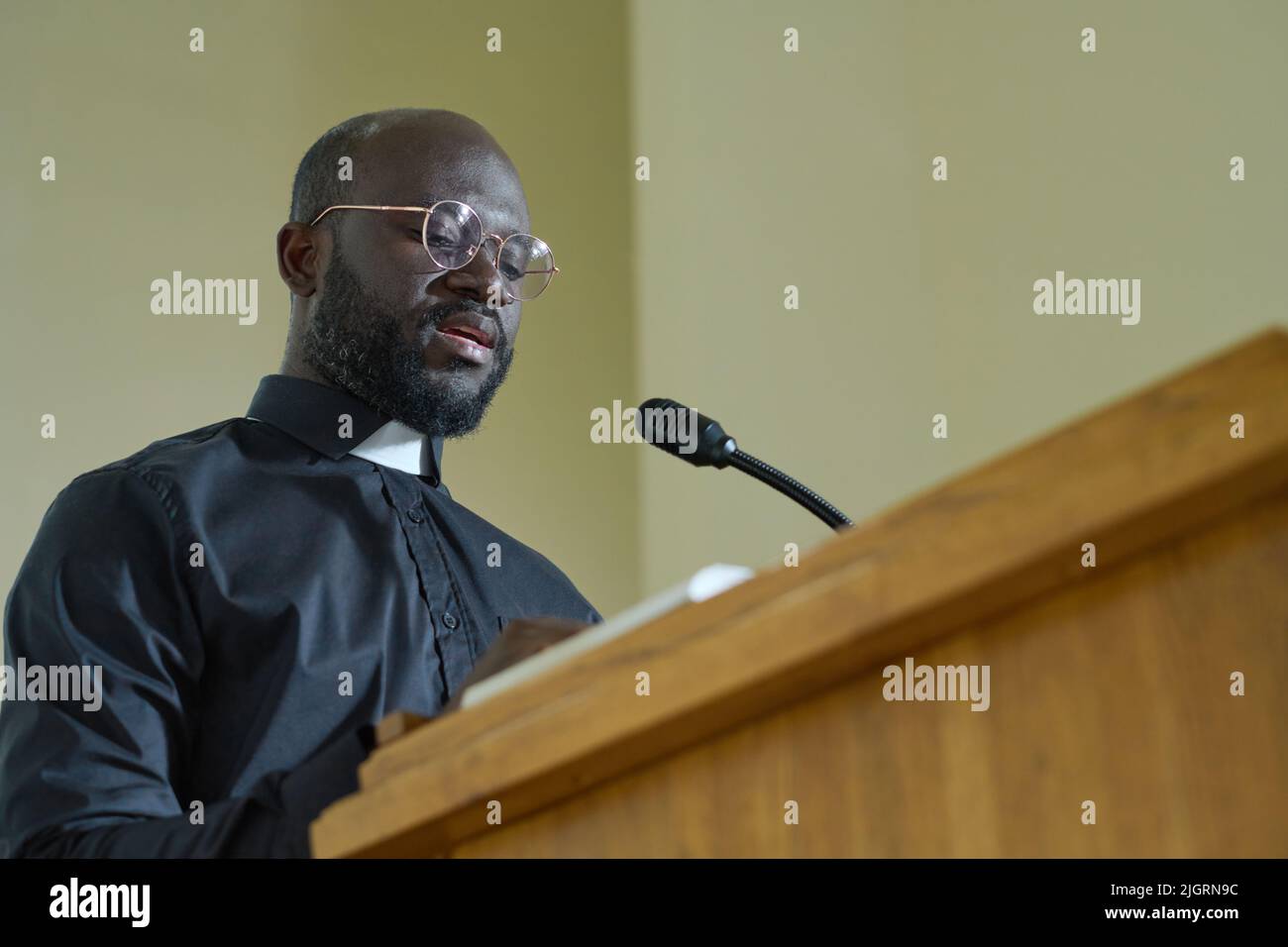 Young priest in black shirt with clerical collar reading verses from Bible and explaining them while pronouncing speech by pulpit Stock Photo