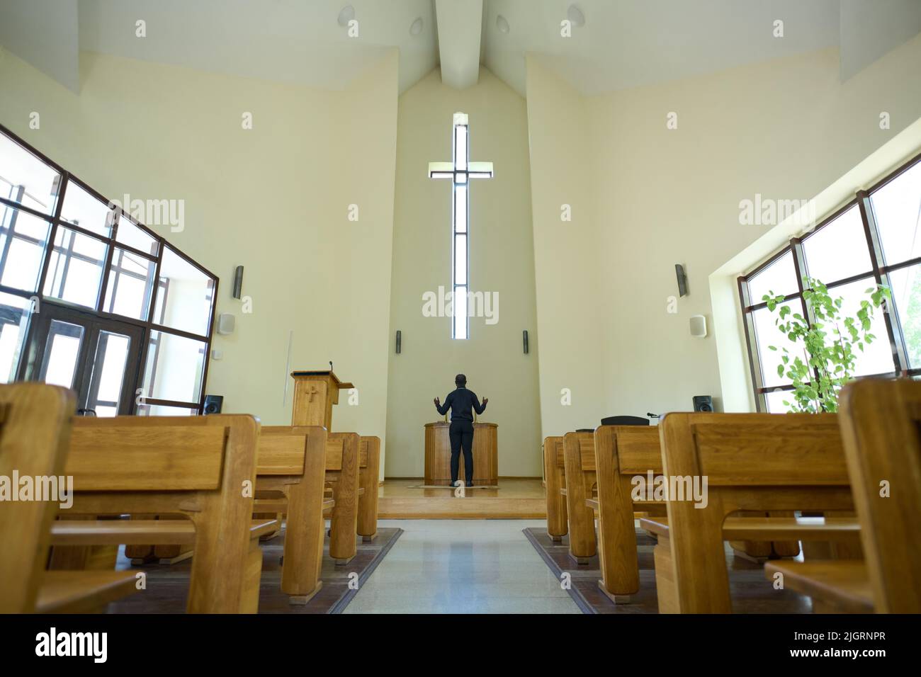 Interior of church with long aisle leading to pastor standing in front of cross and wooden pulpit and keeping his arms in blessing gesture Stock Photo