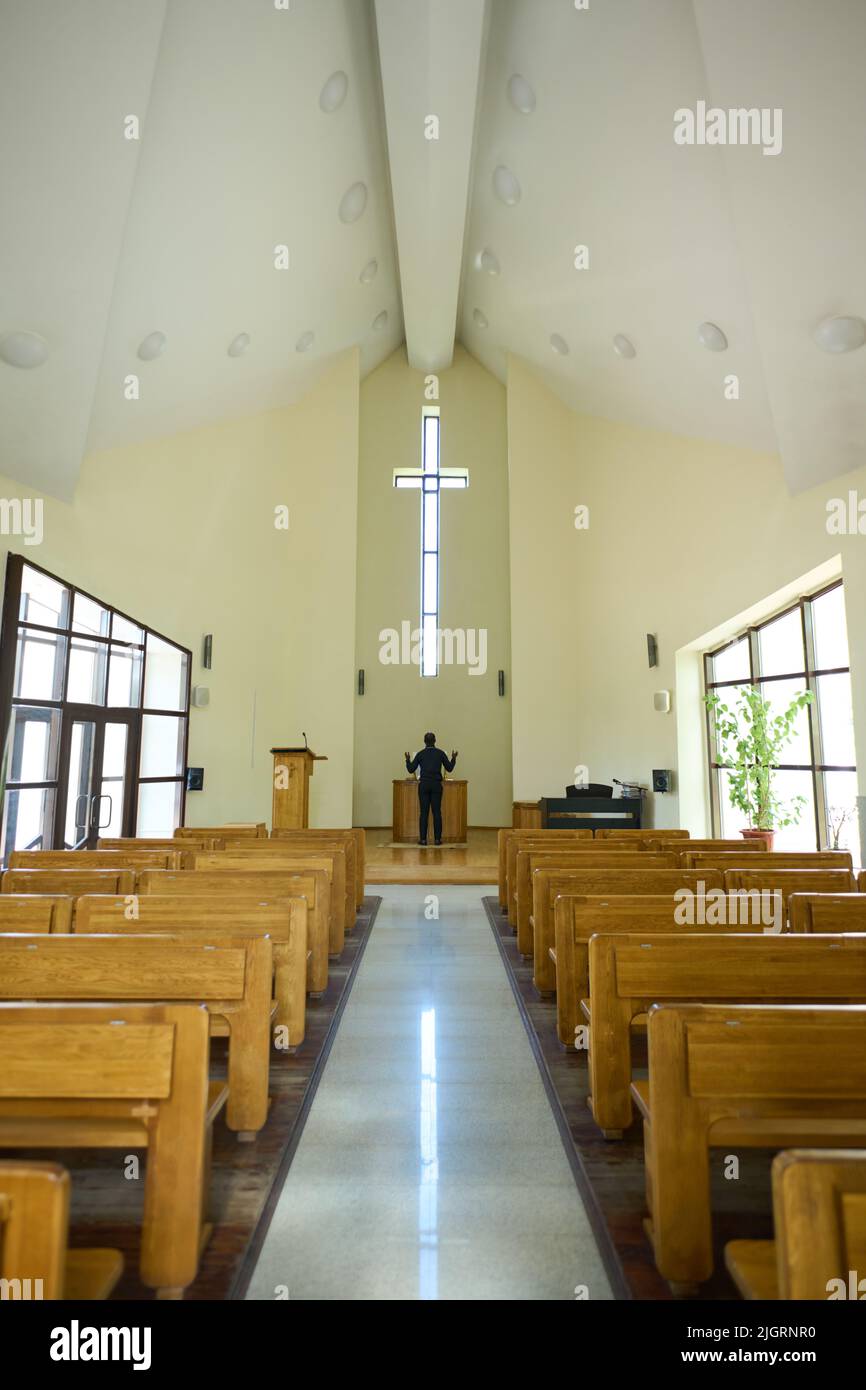 Long aisle between two rows of wooden benches leading towards priest standing in front of altar and looking at cross with his arms raised Stock Photo