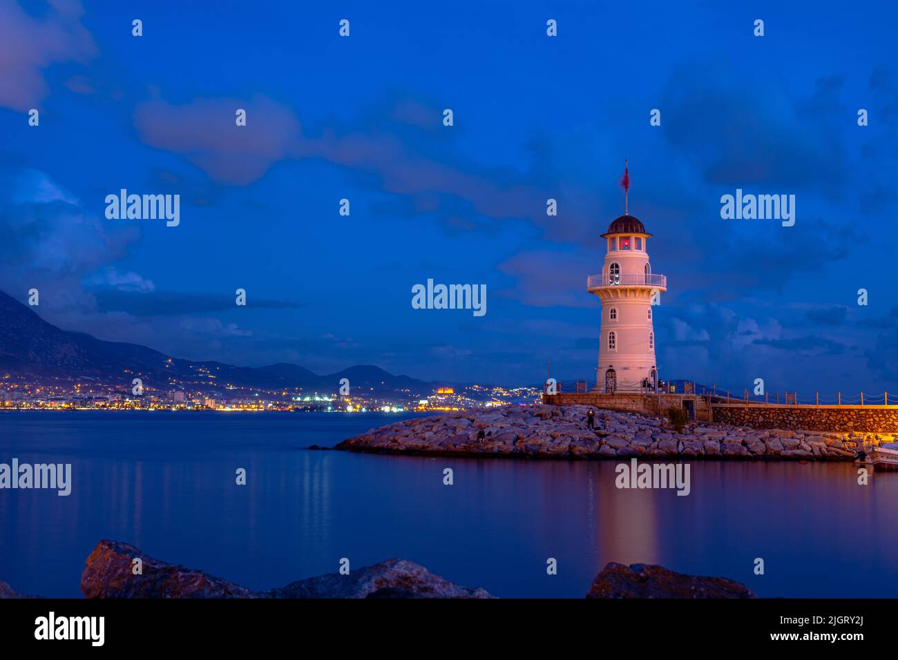 Beautiful night shot of the lighthouse on the pier in the old town of Alanya. City lights and mountains in the background. Long exposure photo. Stock Photo