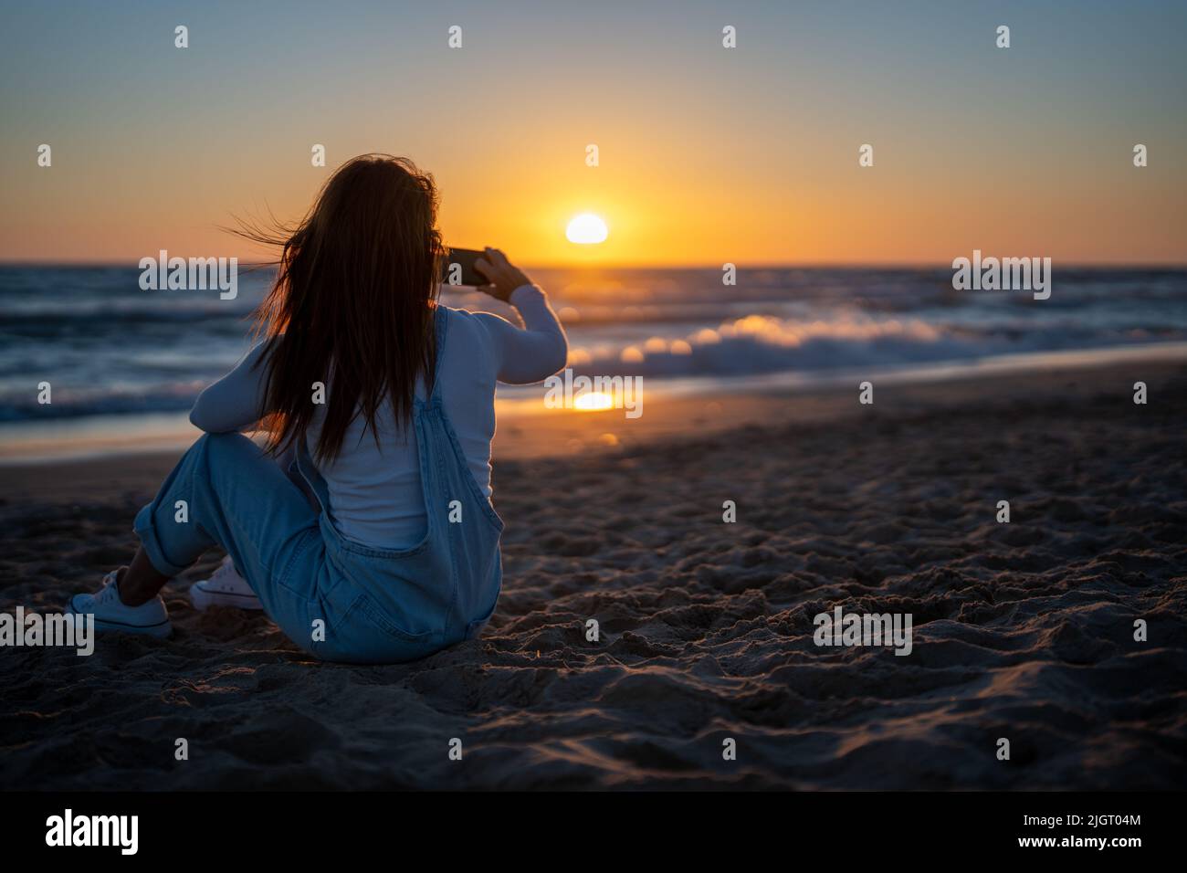 red-haired woman sitting on the sand of a beach shooting a video of a sunset Stock Photo