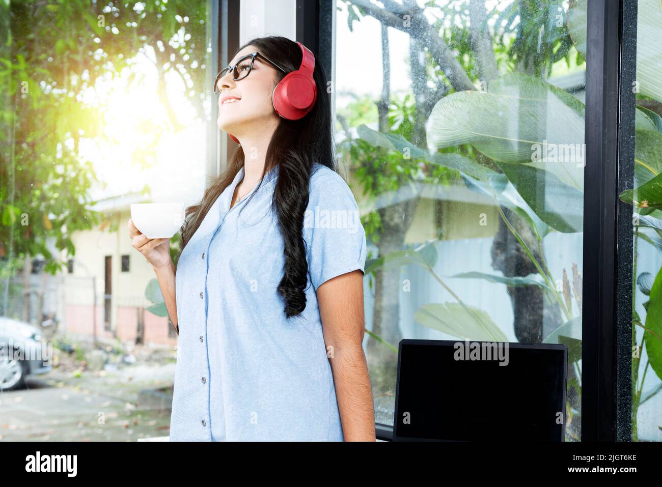 Asian woman with eyeglass listening to music with headphones and holding a cup of coffee with a laptop on the table in the coffee shop Stock Photo