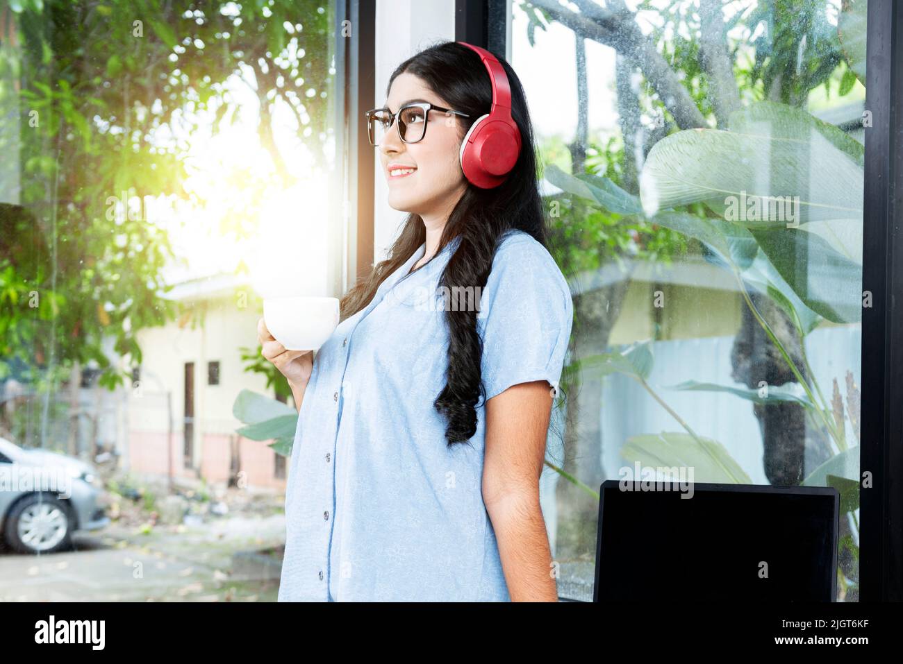 Asian woman with eyeglass listening to music with headphones and holding a cup of coffee with a laptop on the table in the coffee shop Stock Photo