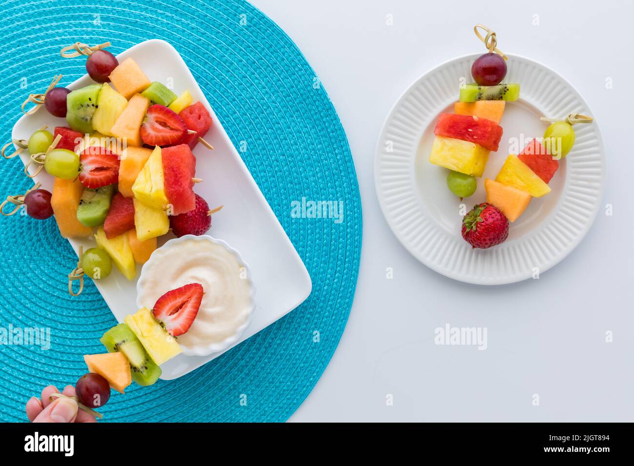 Fresh fruit kebabs on a platter with a hand dipping one into a yogurt