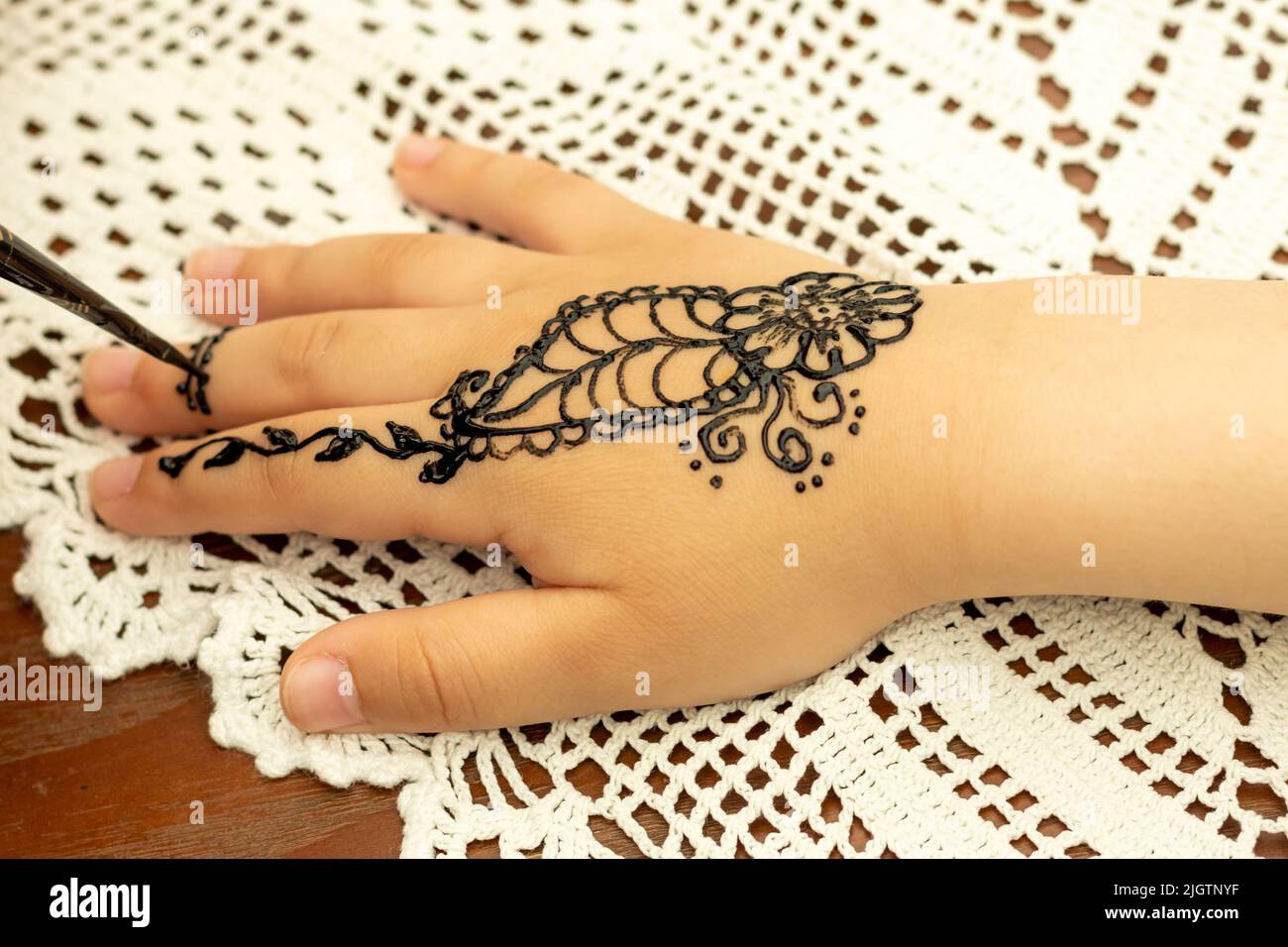 A girl draws henna patterns on a child's hand against a white tablecloth during a school fair. Stock Photo