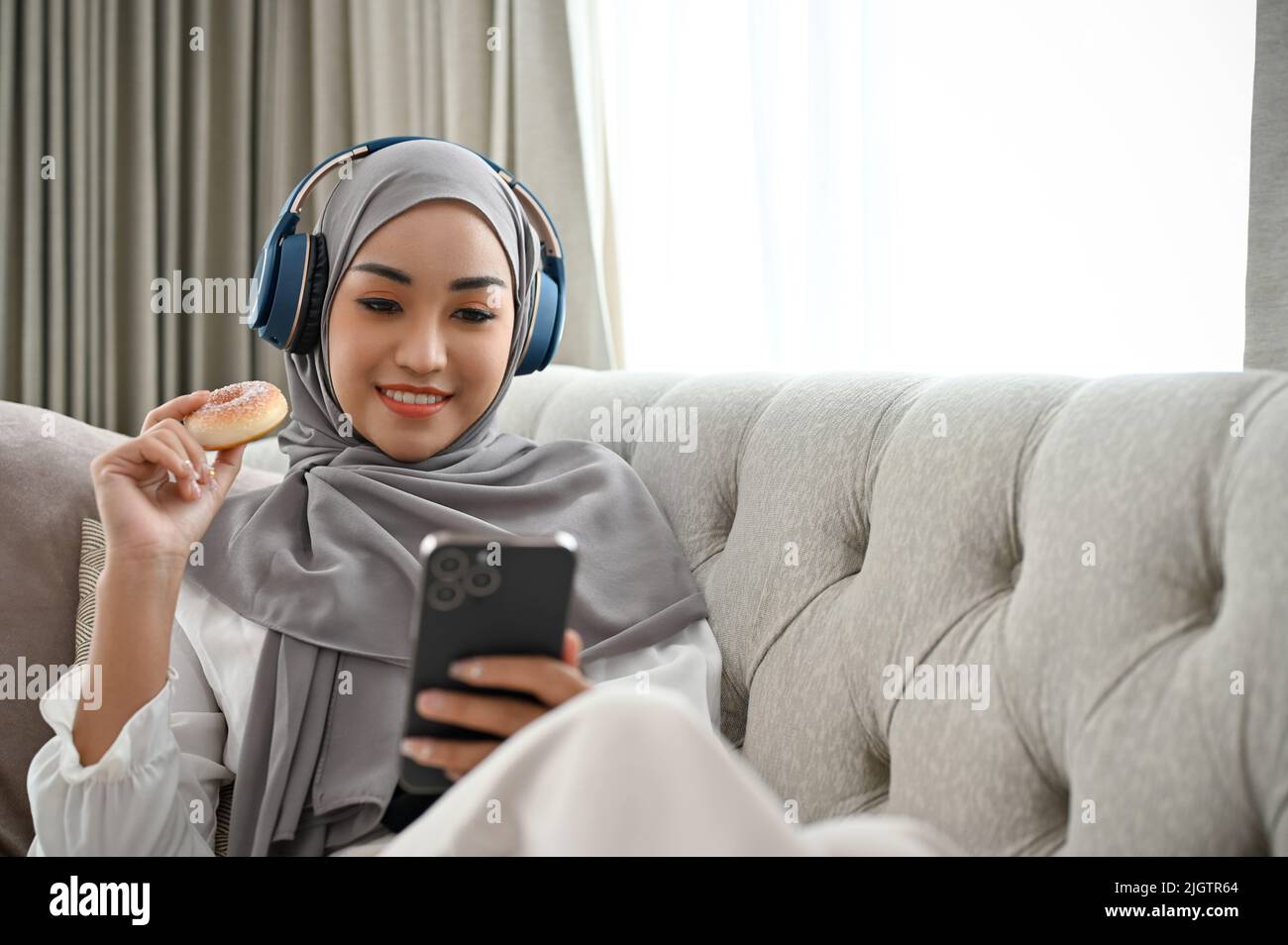 Attractive young Asian Muslim woman wearing hijab and headphones relaxes on the sofa, using her smartphone to listen to music and eat doughnuts. Stock Photo