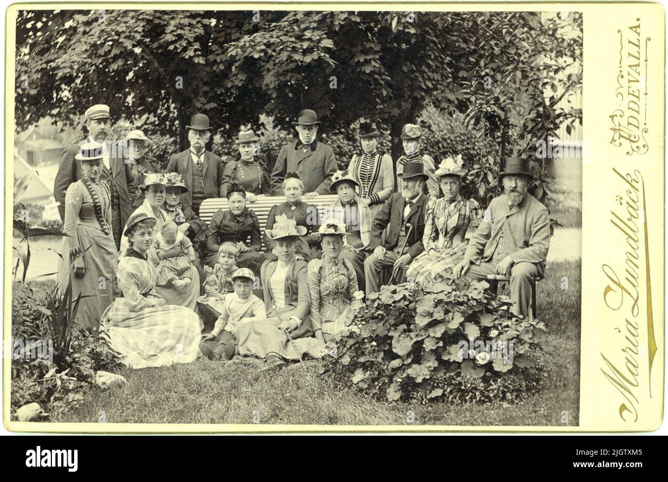 Text in gold on the front of the photo: 'Maria Lundbäck. Uddevalla.' Written text on the back: 'Summer 1891. Mayor Radhe, Mrs. Radhe, Olga Radhe, Elsa Radhe, Anna Radhe, Archbishop Anton Niklas Sundberg, Mrs. Sundberg, Prof. Berglind, Mrs. Berglind, Olga Berglind, Carry (?) Öhman. Rev. secr. Reuterswaard, Mrs. Reuterswaard. ”A group of well -dressed people in a garden. Some sit on a bench, some sit on the ground and some get up. In front of the company you see two discounts. Photographer Maria Lundbäck was born in Uddevalla where her father was a brewery owner. She learned the photography prof Stock Photo