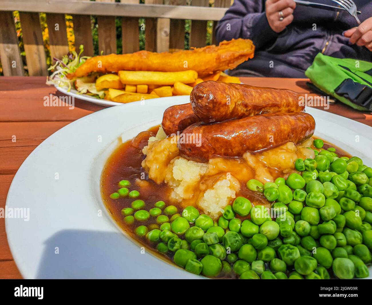 Bangers (Sausages) and Mash with Onion Gravy and peas. Fish and chips in the background at Devonshire Pub, UK Stock Photo