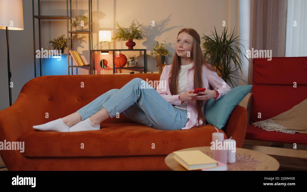 Young teen child girl lying on sofa uses mobile phone smile at modern