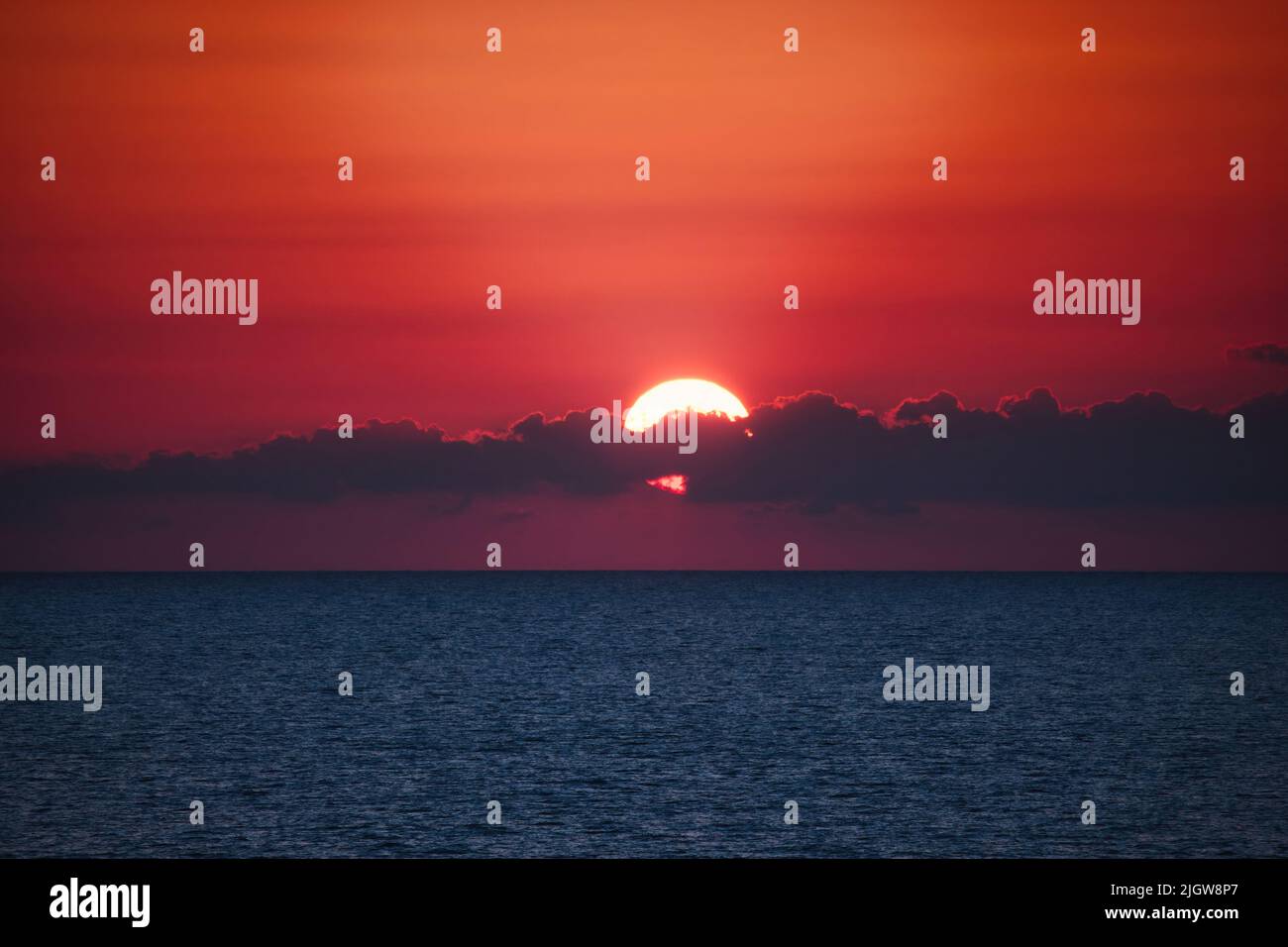 Dramatic sunset over the ocean horizon at dusk with clouds partly covering the sun against a deep red sky background - dreamy glowing light fading int Stock Photo