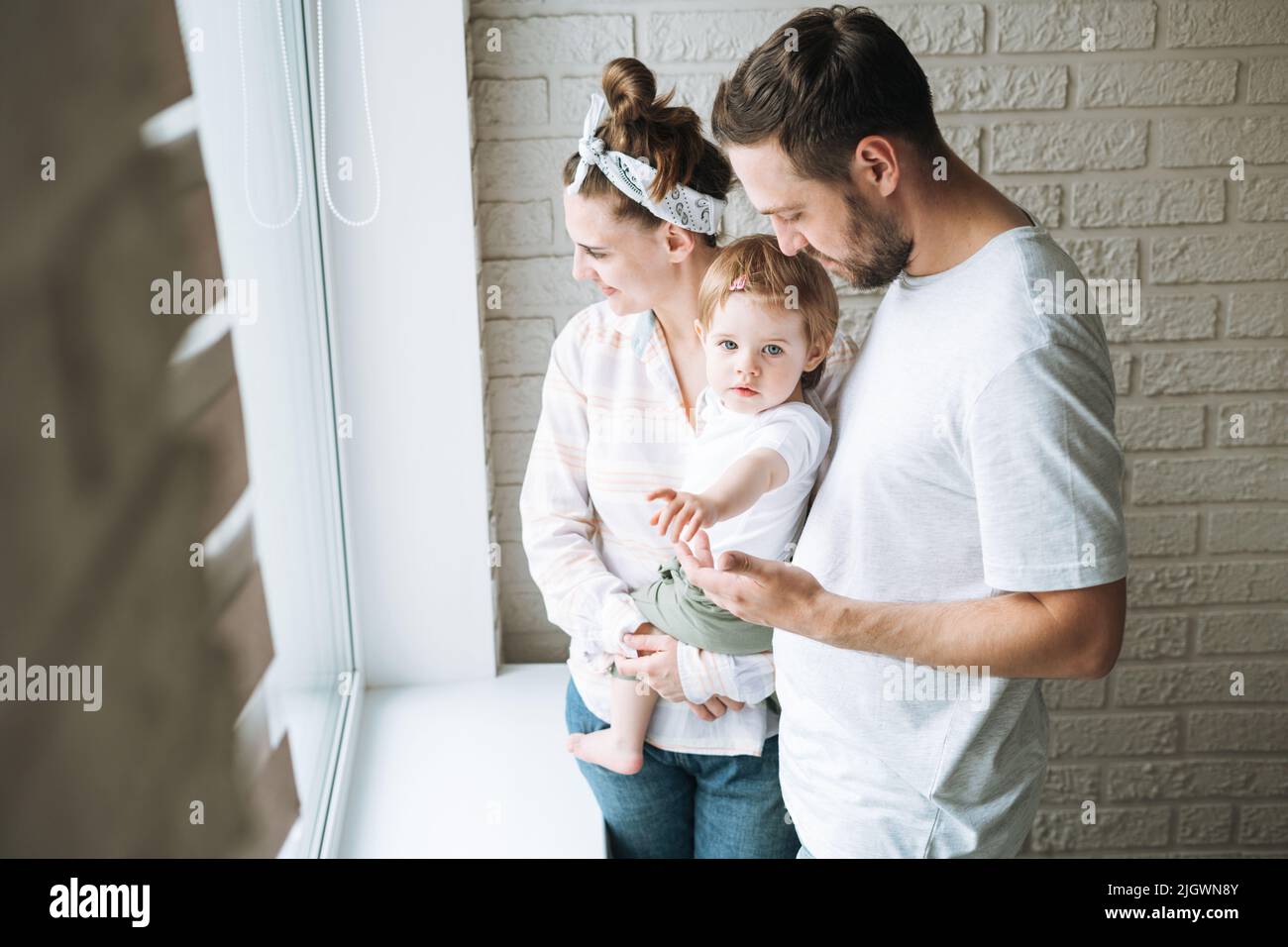 Young family man father and mother woman with baby girl on window sill looking at window at home Stock Photo