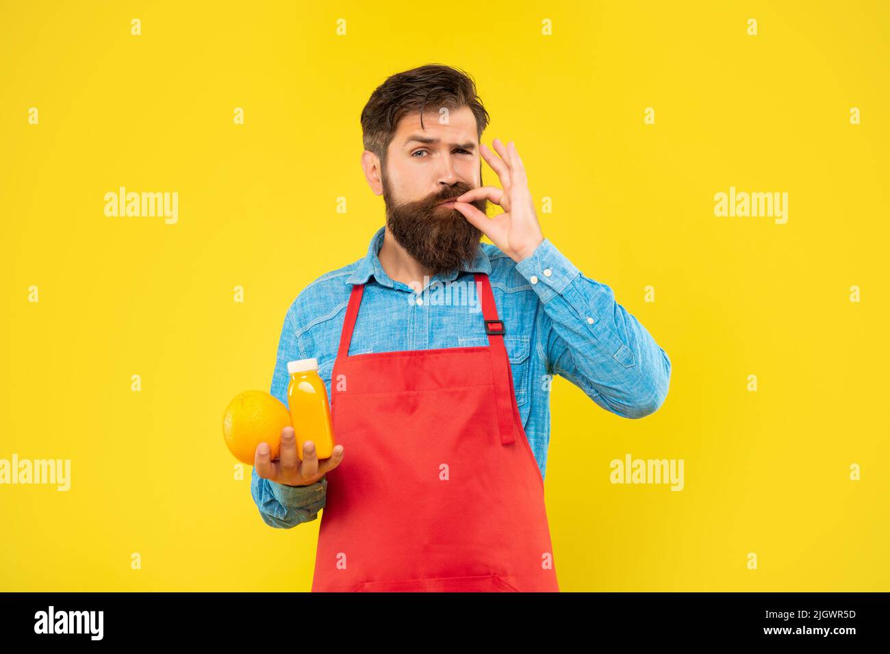Man in apron licking finger holding orange and juice bottle yellow background, juice barkeeper Stock Photo