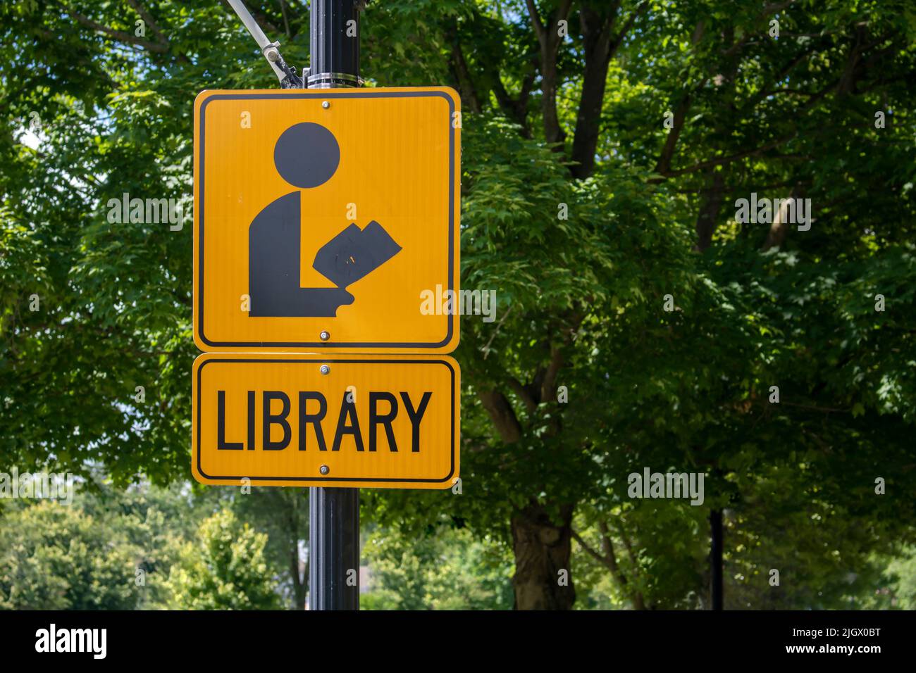 orange and black street and sidewalk sign with an icon of a figure reading a book with the word Library. Stating there's an of the public library near Stock Photo