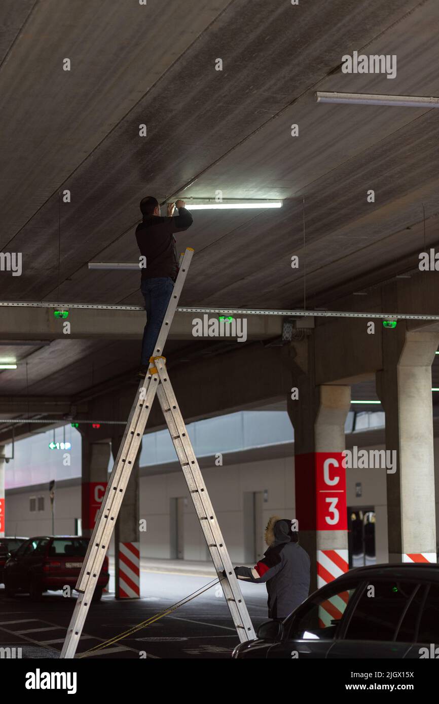City Worker electrician repairs LCD lamp on mall parking. The second worker backs him up. Profession that provides comfort, electricity in cities. Stock Photo
