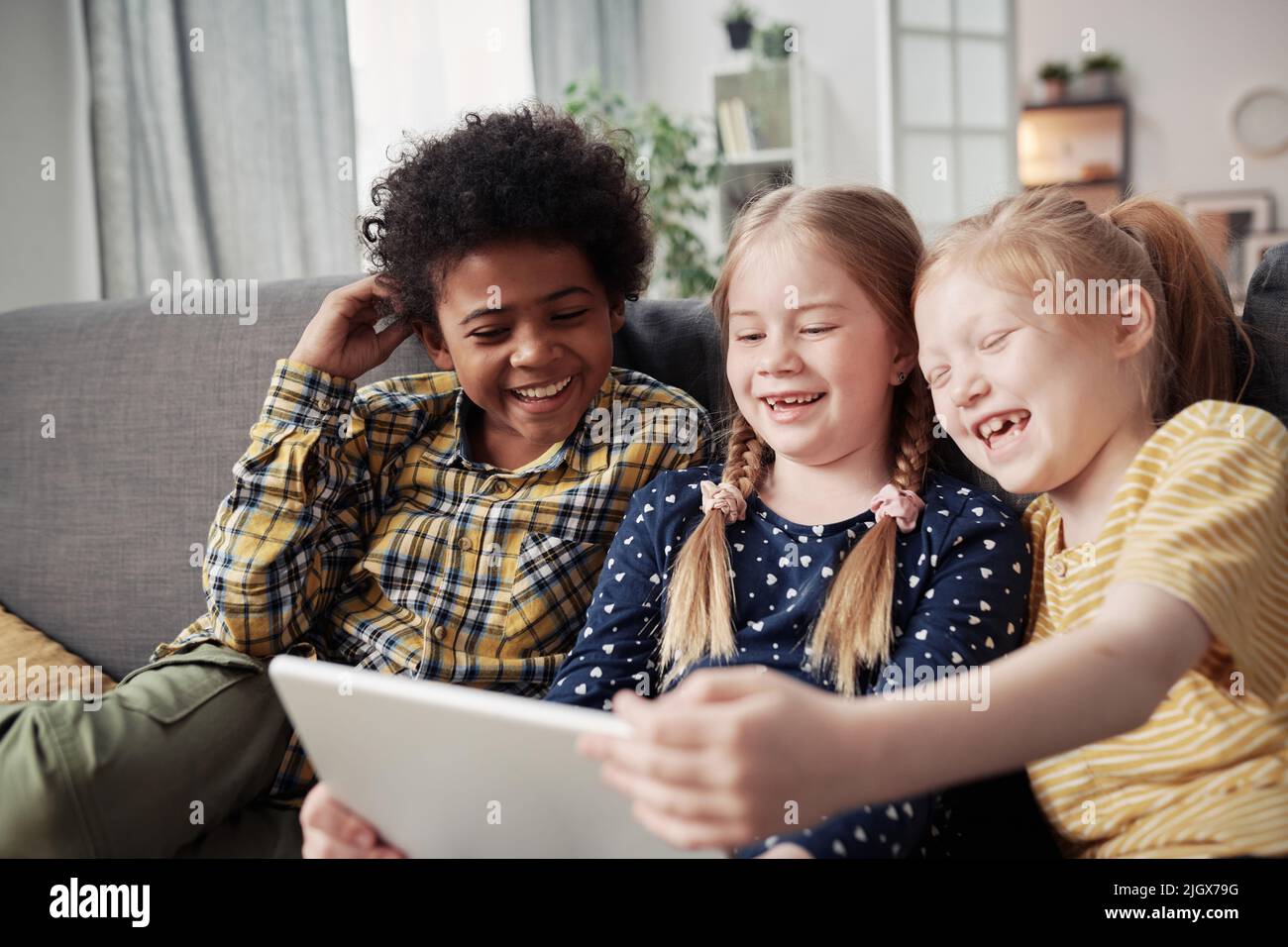 Happy children watching funny video on digital tablet and laughing while sitting together on sofa in the room Stock Photo