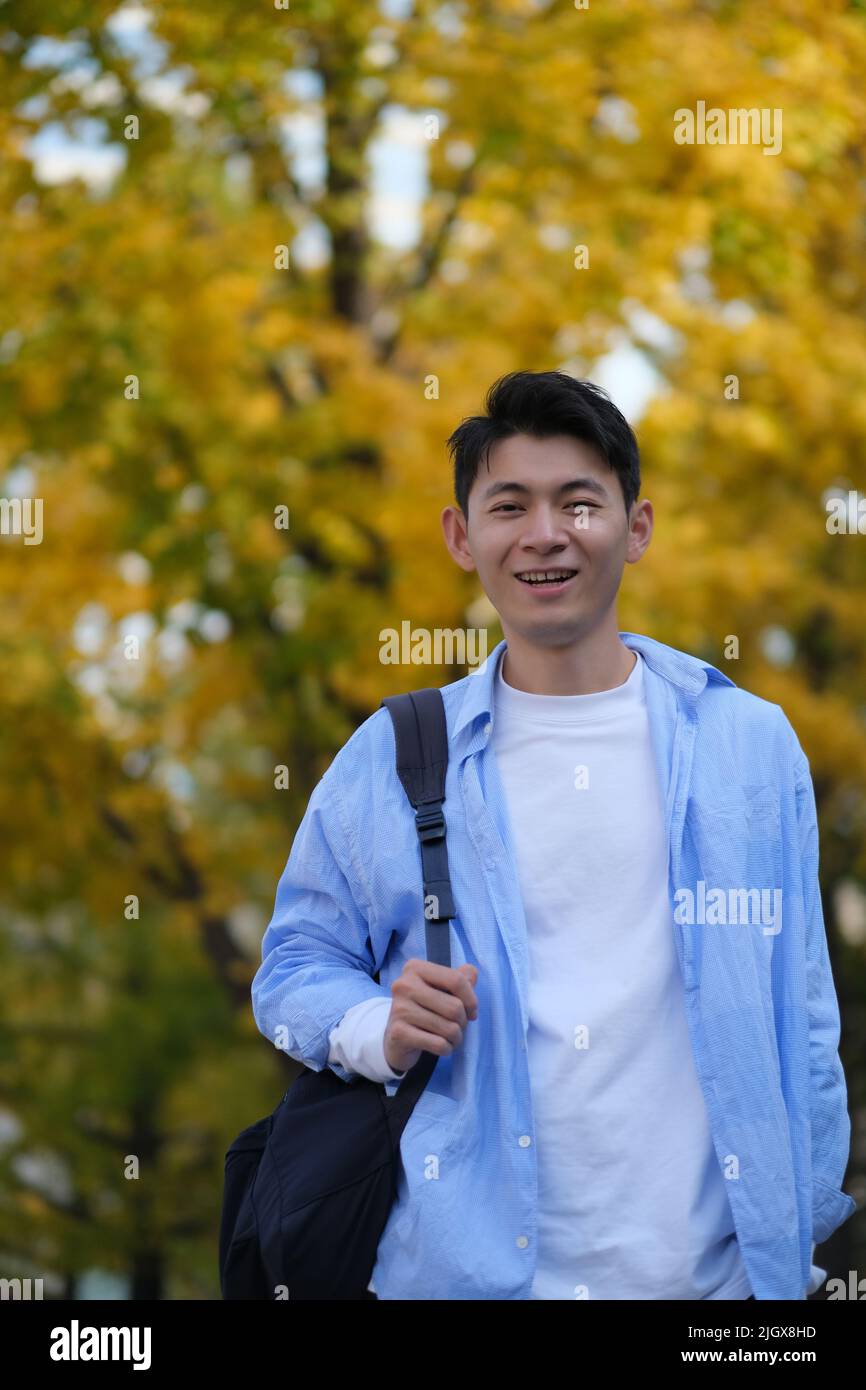 Asian male college student and blurred yellow ginkgo biloba tree in autumn. Carrying a shoulder bag and smiling at camera Stock Photo