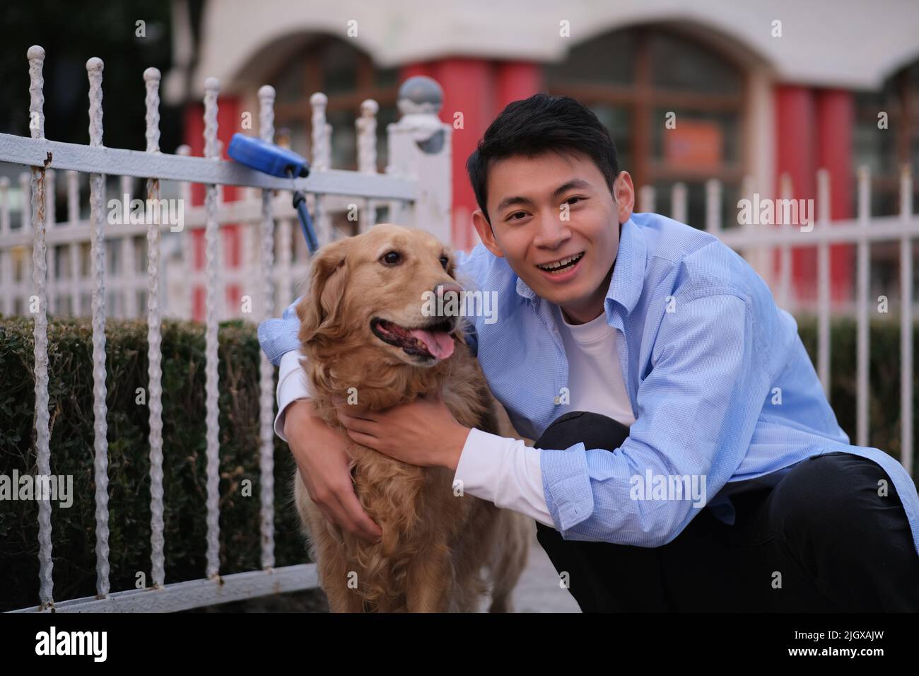 handsome Asian young man cuddle his golden retriever pet dog, smiling at camera in public park Stock Photo