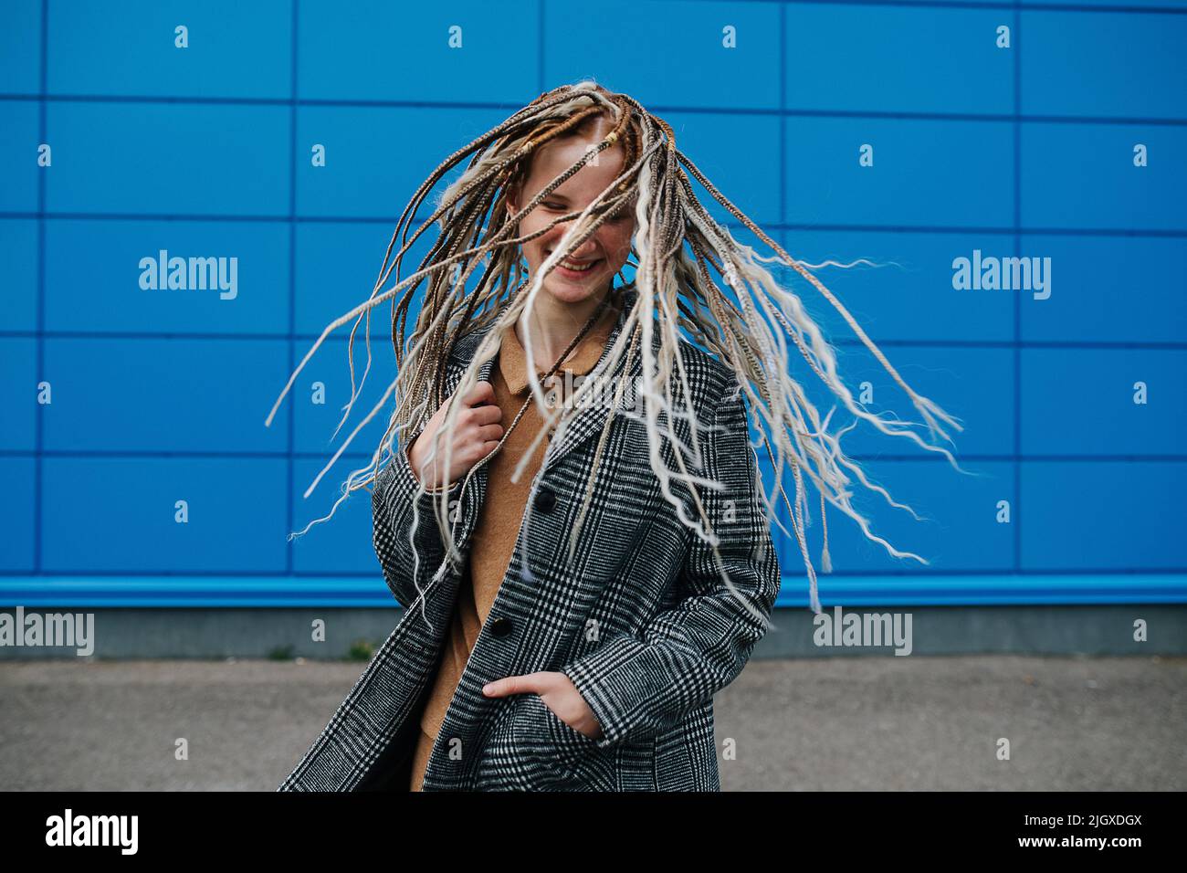 Shaking dreads lighthearted teenage girl in front of a blue panel wall covering. She is wearing a grey checkered jacket. Portrait. Stock Photo