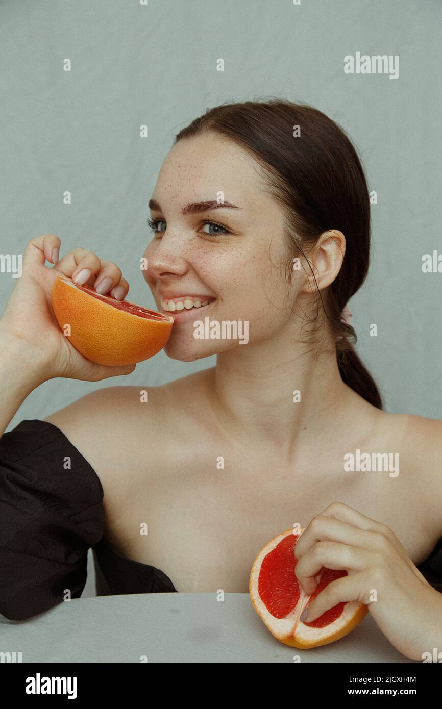 Young woman with freckles playfully bites half of a grapefruit Stock Photo