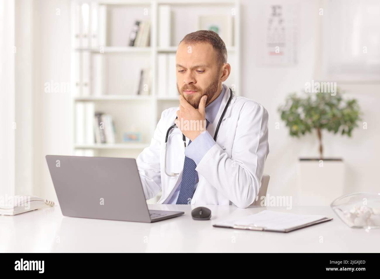 Serious male doctor sitting in an office and looking at a laptop computer Stock Photo