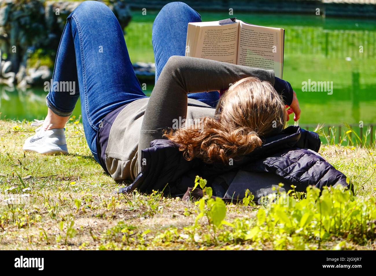 Young American woman, on grass with head propped on jacket, reading a novel, hard backed book. Enjoying a sunny Italian day in April with a good book. Stock Photo