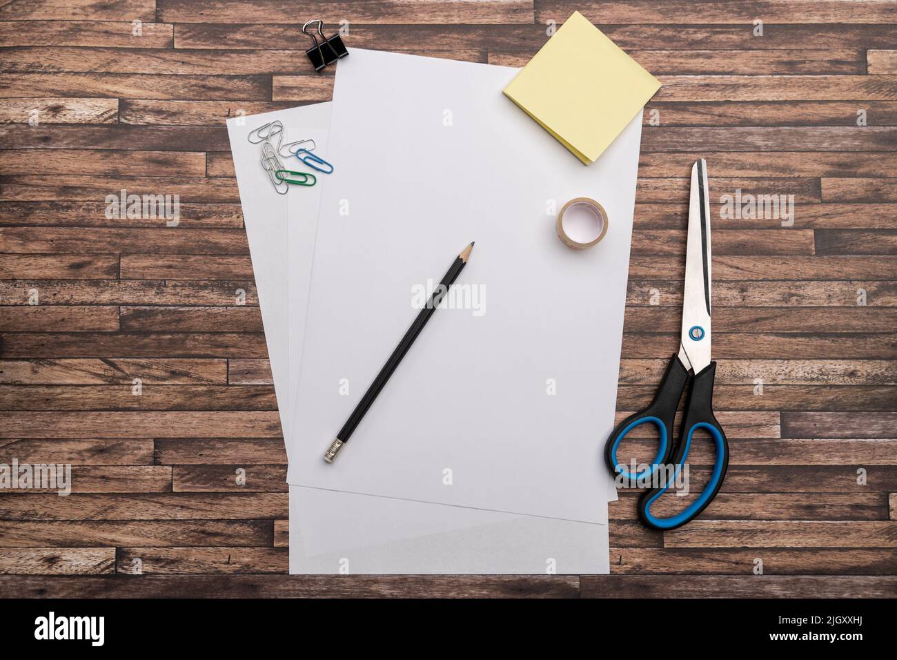 Empty white paper on a desk with office supplies lying down. Top down view from above on a table with a scissors, adhesive tape, a pencil, notes. Stock Photo