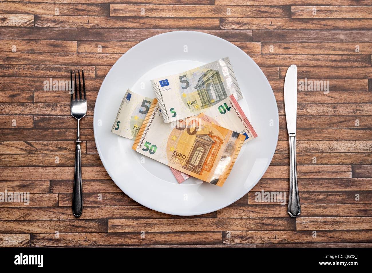 Euro banknotes lying on a dinner plate with cutlery on the table. Symbolic image for eating money notes. Food is getting expensive due to inflation. Stock Photo