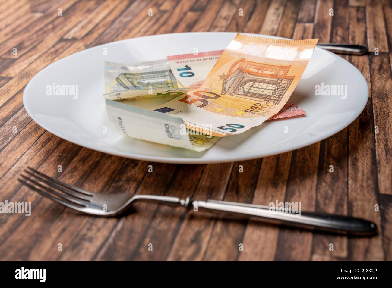 Euro banknotes lying on a dinner plate with cutlery on the table. Symbolic image for eating money notes. Food is getting expensive due to inflation. Stock Photo