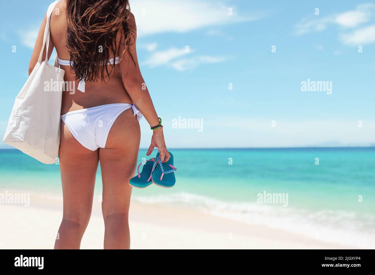 Sexy bikini girl on the beach, young female with swimsuit from behind in empty tropical beach holding flip flops and tote bag with turquoise ocean in Stock Photo