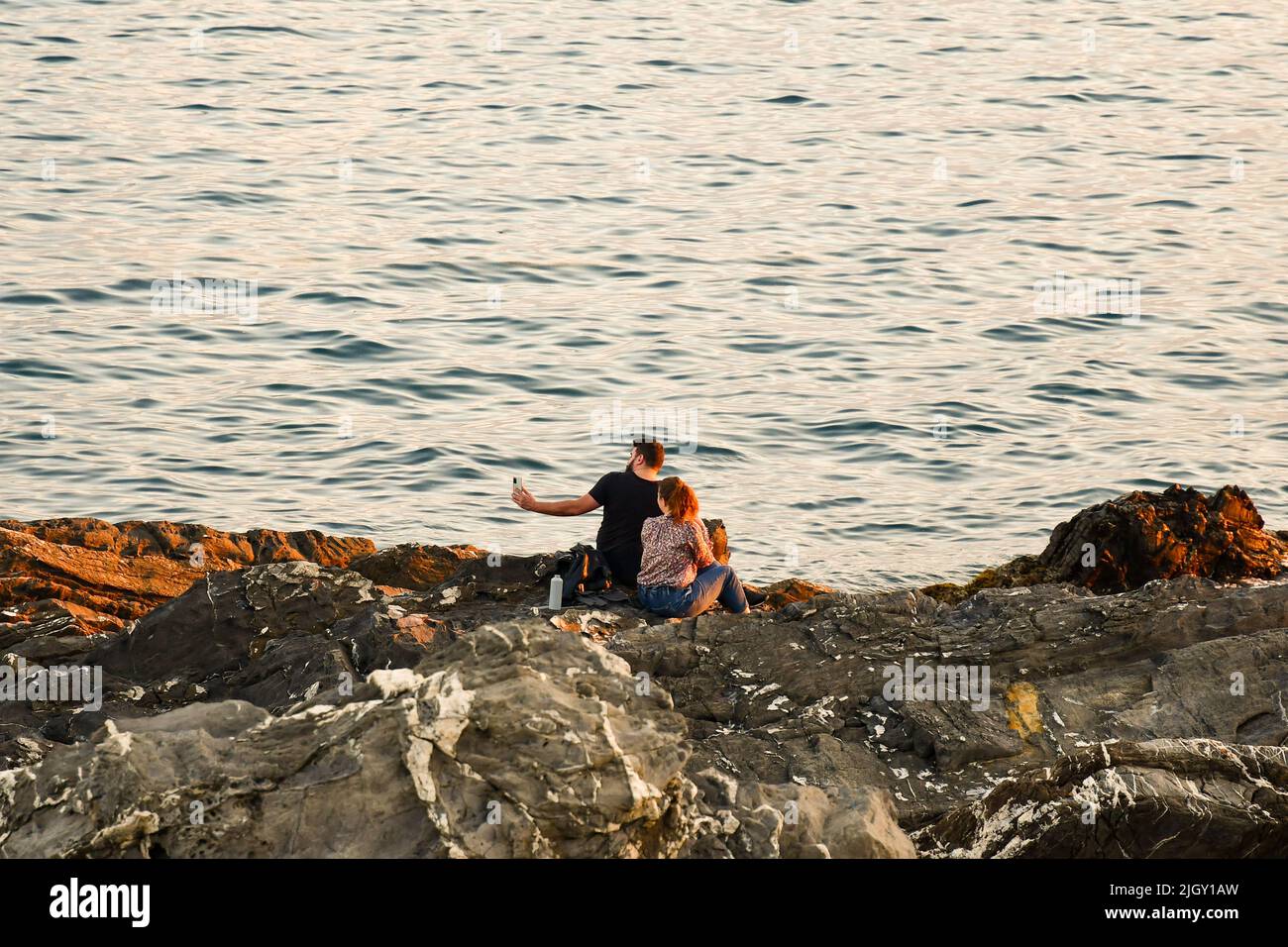 A young couple (about 20 years old) sitting on the cliff by the sea and taking selfies with a smartphone at sunset, Nervi, Genoa, Liguria, Italy Stock Photo