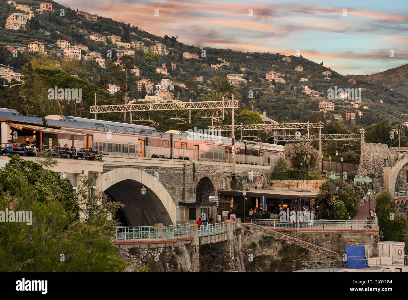 A stretch of the Anita Garibaldi Promenade with people at outdoor cafe and a train in the railway station at sunset, Nervi, Genoa, Liguria Stock Photo
