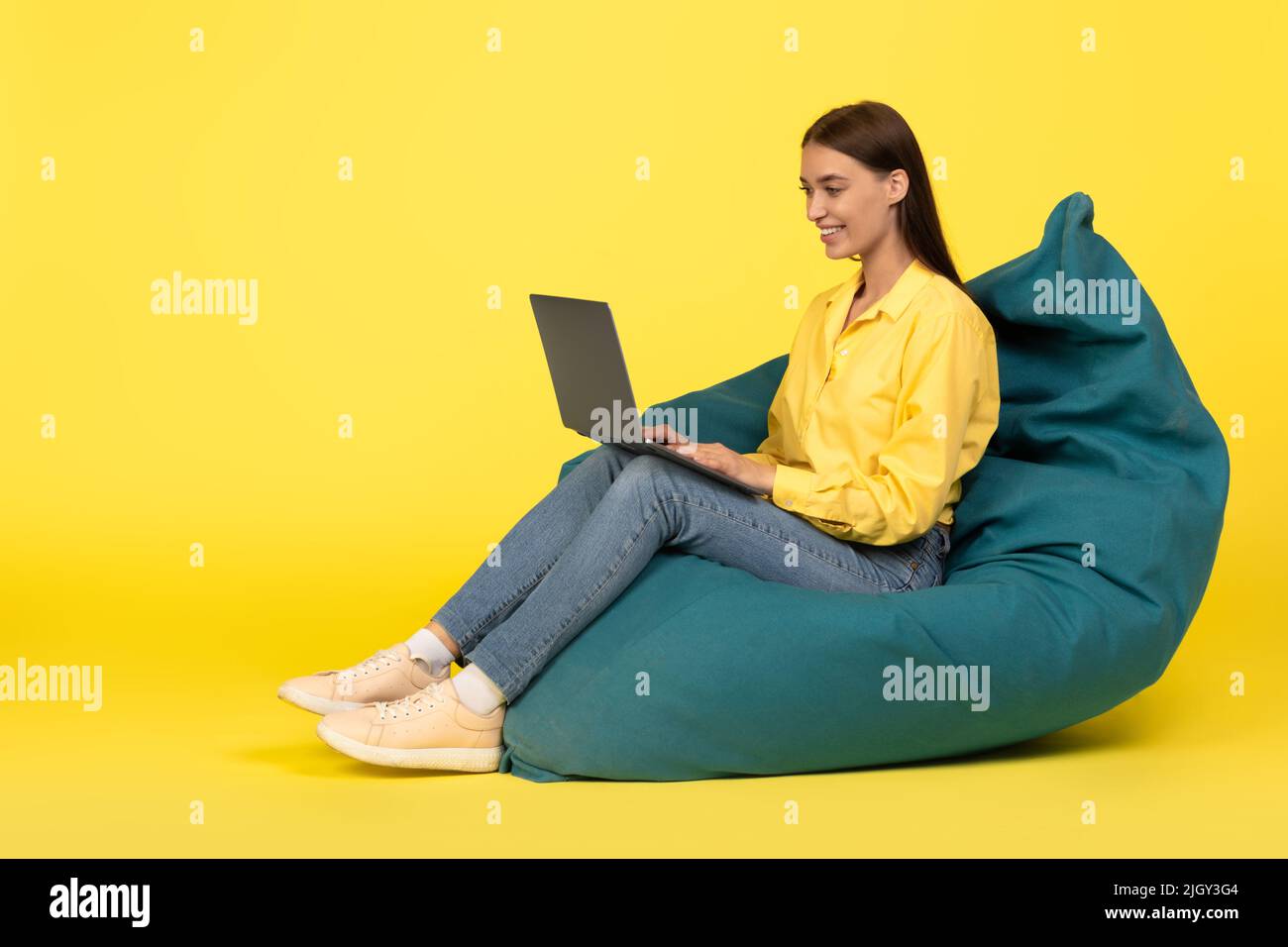 Woman Using Laptop Sitting In Beanbag Chair Over Yellow Background Stock Photo