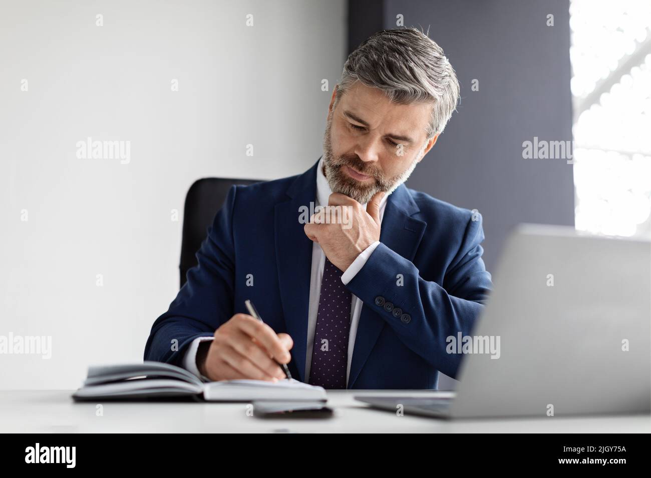 Busy Handsome Middle Aged Businessman Writing In Notepad While Sitting In Office Stock Photo